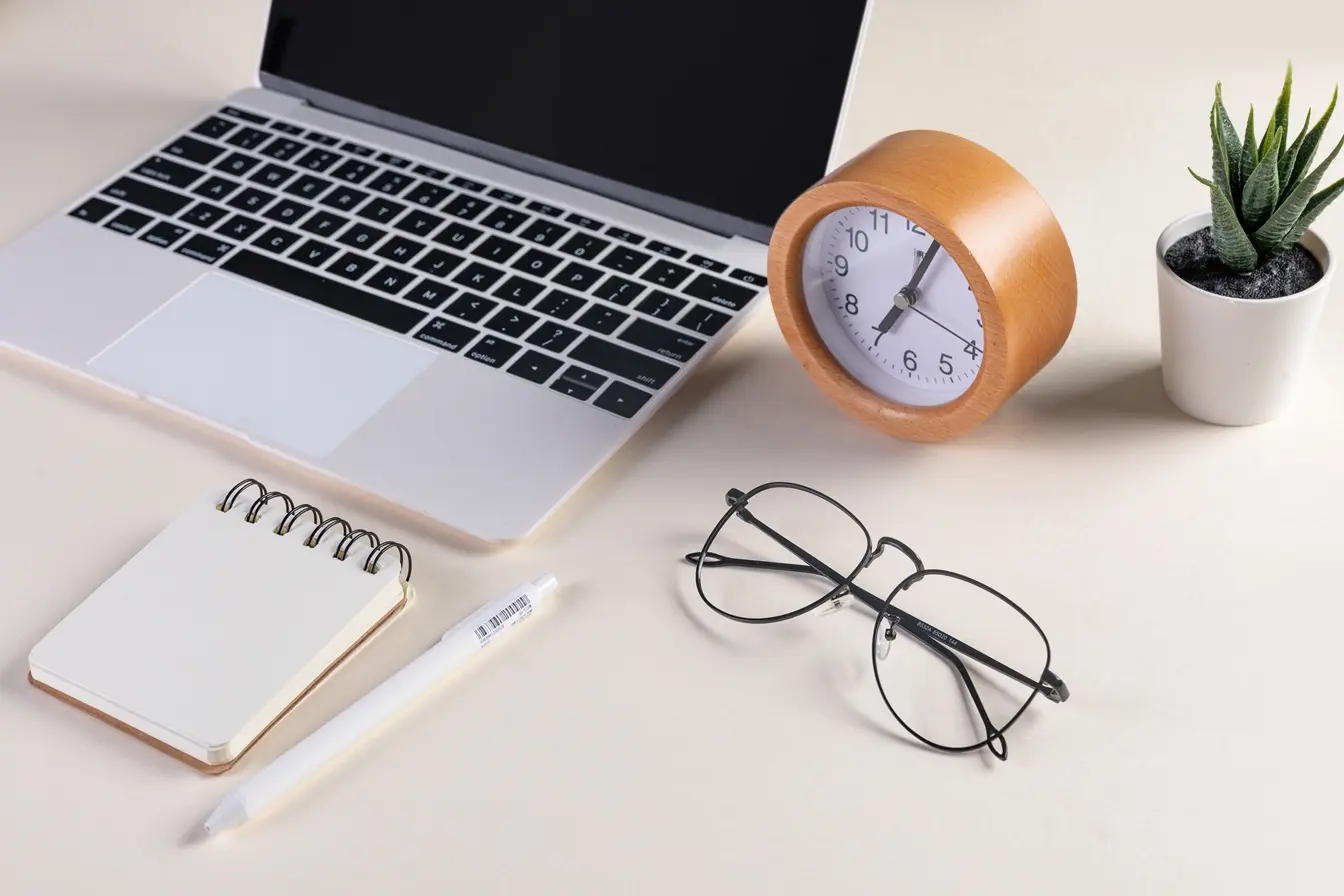 Bureau épuré avec ordinateur portable, horloge en bois, carnet et lunettes posés avec soin, illustrant une organisation de travail claire et sereine.