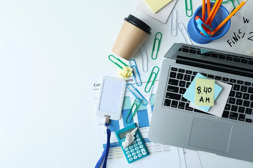 Vue de dessus d'un bureau encombré avec ordinateur portable, trombones épars, post-its et calculatrice, illustrant le désordre d'une organisation bureautique non structurée.